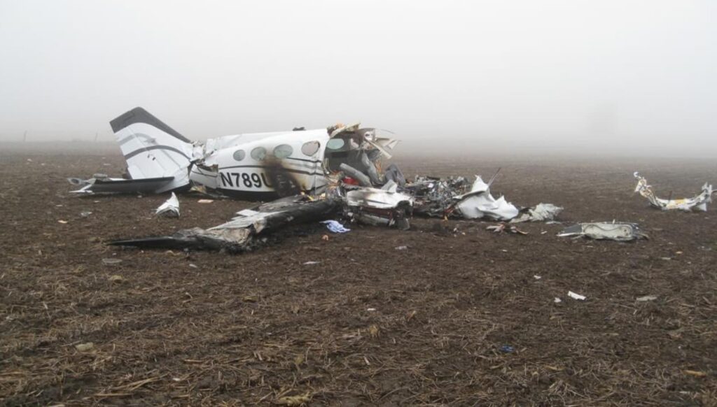 Right side view of the wrecked Cessna 414A with tail number N789UP visible, surrounded by scattered debris in a fog-covered farm field.